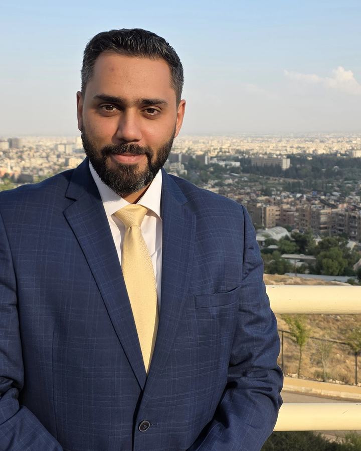 Dr. Sari Hamoud in a navy checked suit and gold tie, with a city panorama behind him