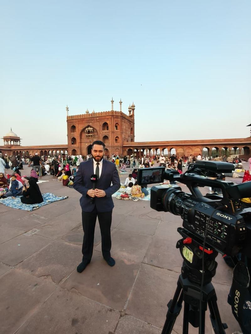 Dr. Sari Hamoud standing in the courtyard of Jama Masjid, Delhi, next to a camera on a tripod at dusk