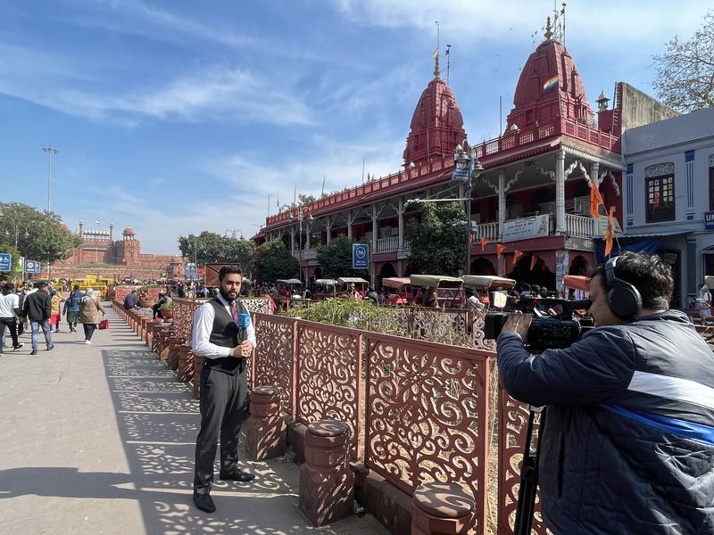 Dr. Sari Hamoud presenting to camera on a pedestrian street in Old Delhi, with the Red Fort behind and a cameraman filming