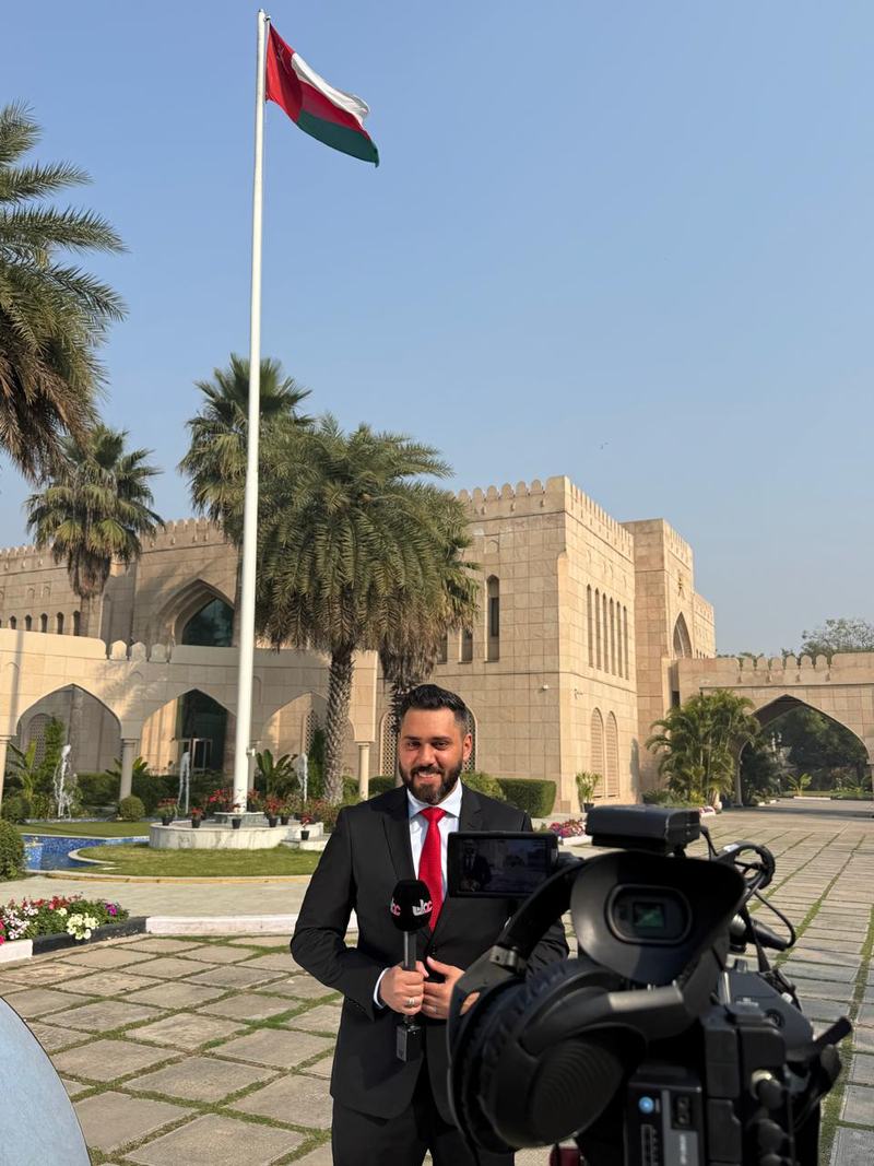 Dr. Sari Hamoud holding a microphone beside a broadcast camera in front of a stone building and a national flag