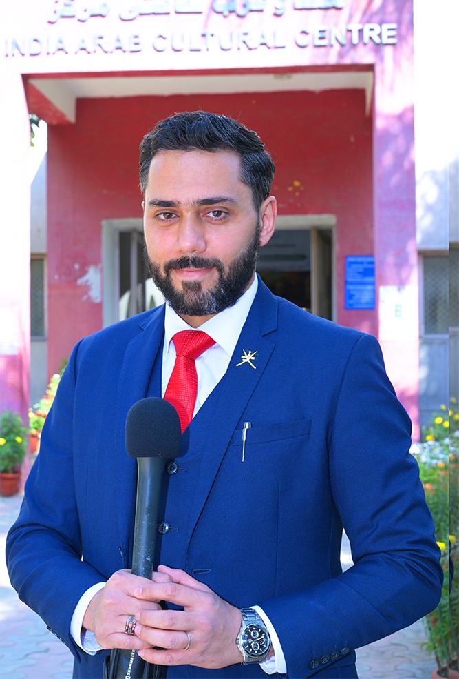 Dr. Sari Hamoud in a blue suit and red tie holding a microphone in front of the India Arab Cultural Centre