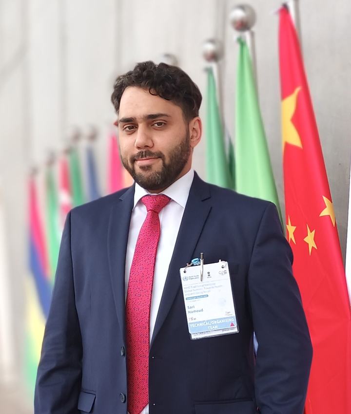 Dr. Sari Hamoud in a navy suit and red tie, wearing a WHO Traditional Medicine Global Summit badge, in front of national flags
